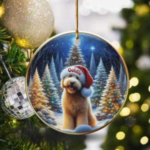 May include: Round Christmas ornament featuring a golden doodle wearing a Santa hat with the name "Goldie". The background shows a snowy forest scene with lit Christmas trees. The ornament is hanging on a Christmas tree.