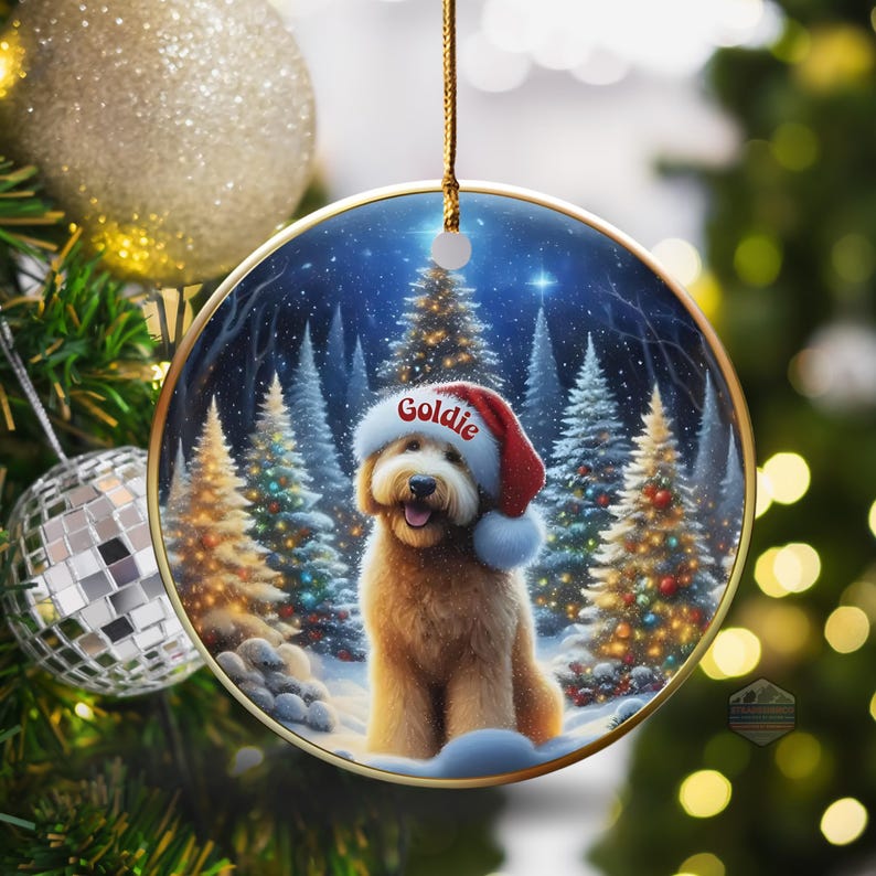 May include: Round Christmas ornament featuring a golden doodle wearing a Santa hat with the name "Goldie". The background shows a snowy forest scene with lit Christmas trees. The ornament is hanging on a Christmas tree.
