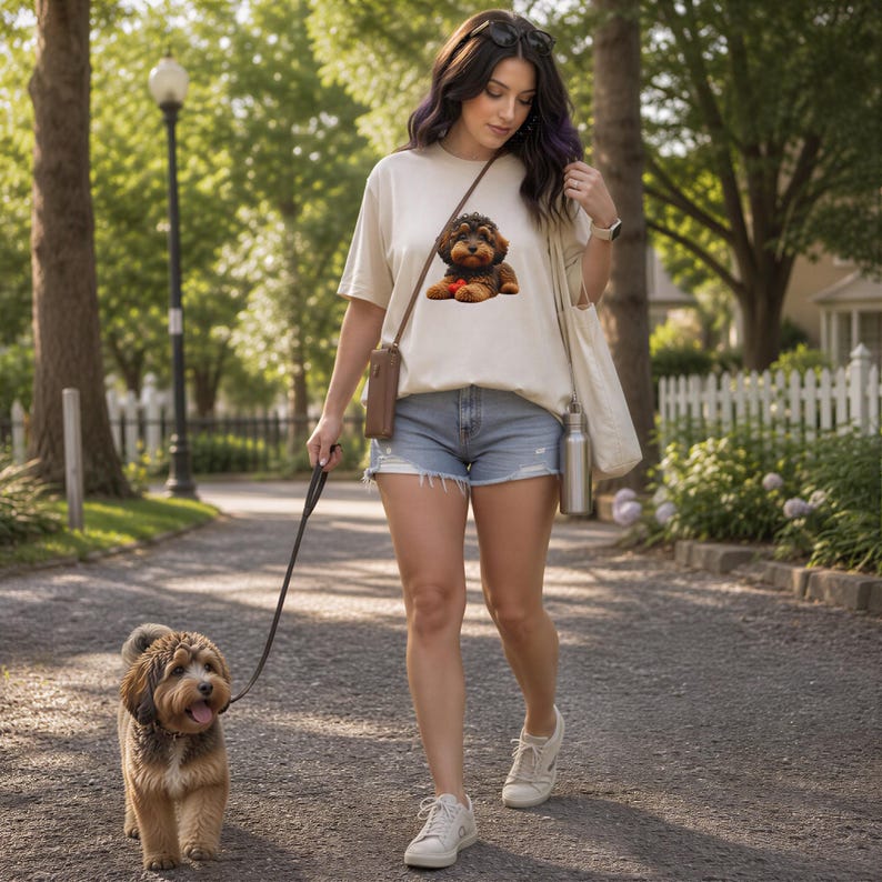 A woman wearing a white t-shirt with a dog graphic walks her small brown and white dog on a leash in a park.