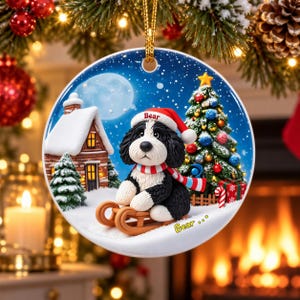 A decorative ornament featuring a black and white dog wearing a Santa hat and riding a sled, with a snowy village scene and a Christmas tree in the background.