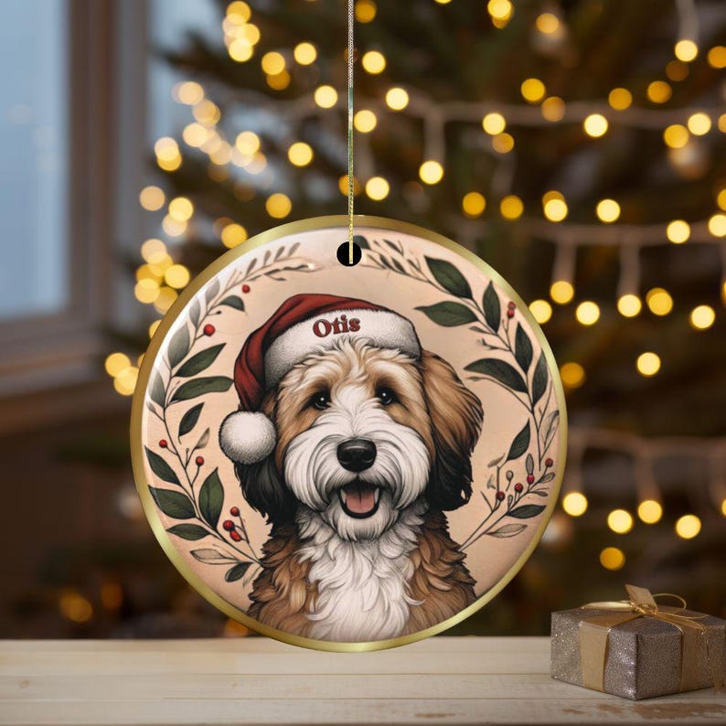 Puede incluir: Un adorno redondo de cer&aacute;mica con un perro marr&oacute;n y blanco que lleva un gorro de Pap&aacute; Noel rojo con el nombre Otis impreso en el borde. El perro est&aacute; centrado dentro de un marco circular de hojas verdes y bayas rojas, colgado de un hilo dorado.