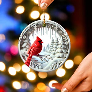 Peut inclure: Ornement de No&euml;l rond et blanc repr&eacute;sentant un cardinal rouge perch&eacute; sur une branche enneig&eacute;e. L'ornement pr&eacute;sente une sc&egrave;ne hivernale avec des arbres enneig&eacute;s et des flocons de neige. L'ornement est tenu par une main avec du vernis &agrave; ongles paillet&eacute;.
