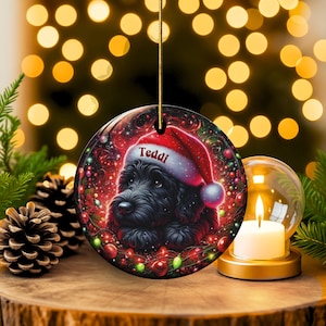May include: A round Christmas ornament featuring a black dog wearing a Santa hat with the name "Teddi" on it. The ornament has a festive design with red and green accents, and a gold string for hanging.