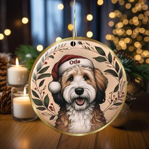 Puede incluir: Un adorno navide&ntilde;o redondo de cer&aacute;mica con un perro marr&oacute;n y blanco que lleva un gorro de Pap&aacute; Noel rojo con el nombre Otis impreso en el borde. El perro est&aacute; enmarcado por una corona de hojas verdes y bayas rojas sobre un fondo beige claro.