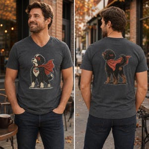May include: A charcoal grey V-neck t-shirt featuring a graphic of a black and white dog wearing a red superhero cape and mask. The front graphic shows the dog with a heart-shaped American flag patch, while the back graphic shows the dog from behind.
