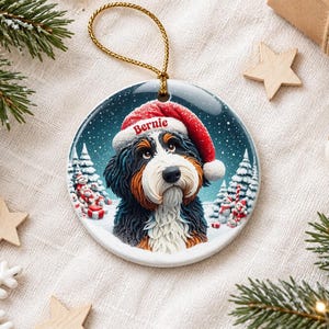 May include: A round ceramic Christmas ornament featuring a Bernese Mountain Dog wearing a red Santa hat with the name Bernie printed on the brim. The dog is set against a snowy winter landscape with pine trees and small gift boxes.