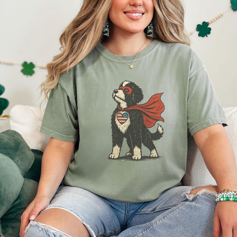 May include: A sage green short-sleeve t-shirt featuring a graphic of a black and white dog wearing a red superhero mask and cape. The dog has a heart-shaped patch on its chest with a red, white, and blue American flag pattern.