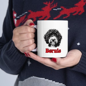 May include: A white ceramic mug featuring a detailed illustration of a black and white Bernedoodle dog face. Below the dog illustration, the name Bernie is printed in bold, red, rounded letters. The mug has a smooth finish and a curved handle.