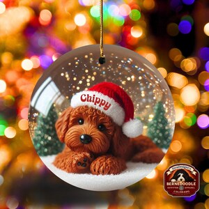 May include: A clear glass Christmas bauble featuring a brown dog wearing a red Santa hat with the name "Chippy" on it. The bauble has a snowy base with small green trees and a gold string for hanging. The background is blurred with bokeh lights.