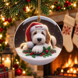 May include: A round ceramic Christmas ornament featuring a fluffy white and tan dog wearing a red Santa hat with the name Bernie printed on the brim. The dog rests on a snowy surface decorated with green holly leaves and red berries.
