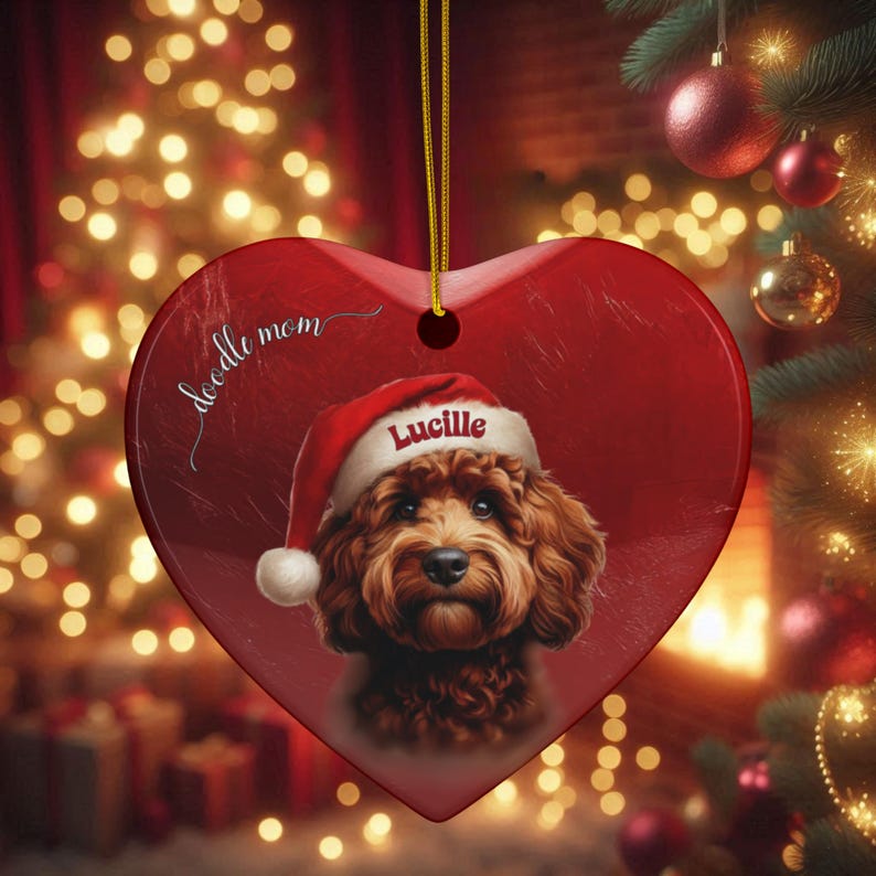 May include: A heart-shaped Christmas ornament with a red background and a portrait of a brown dog wearing a Santa hat. The ornament has the text "doodle mom" and the name "Lucille" on the hat. A gold string is attached for hanging.