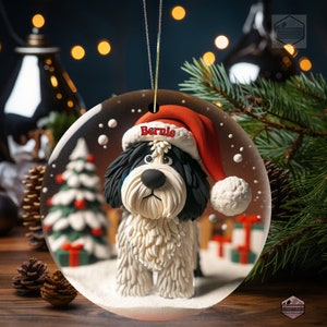 May include: A round ceramic Christmas ornament featuring a black and white dog wearing a red Santa hat with the name Bernie printed in red on the brim. The dog is depicted against a snowy background with a small Christmas tree and presents.