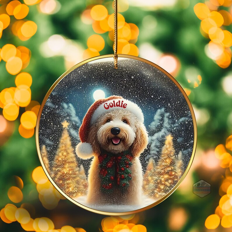 May include: A round Christmas ornament with a gold rim, featuring a fluffy dog wearing a Santa hat with the name "Goldie". The dog is also wearing a festive garland. The background shows a snowy winter scene with Christmas trees.