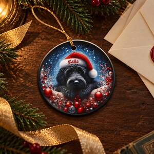 May include: A round ceramic Christmas ornament featuring a black dog wearing a red and white Santa hat with the name Stella printed on the brim. The dog is surrounded by red berries and festive foliage against a dark, starry background.