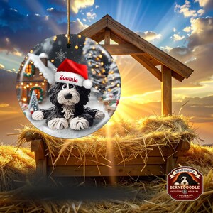 May include: A round Christmas ornament featuring a black and white Bernedoodle dog wearing a Santa hat with the name "Zoomie". The ornament has a winter scene background with a house and Christmas tree. A gold string is attached for hanging.