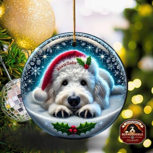 May include: A round Christmas ornament featuring a white Bernedoodle dog wearing a red Santa hat with the word "Snowflake". The ornament has a snowy background with holly and berries. The ornament is hanging from a gold string.