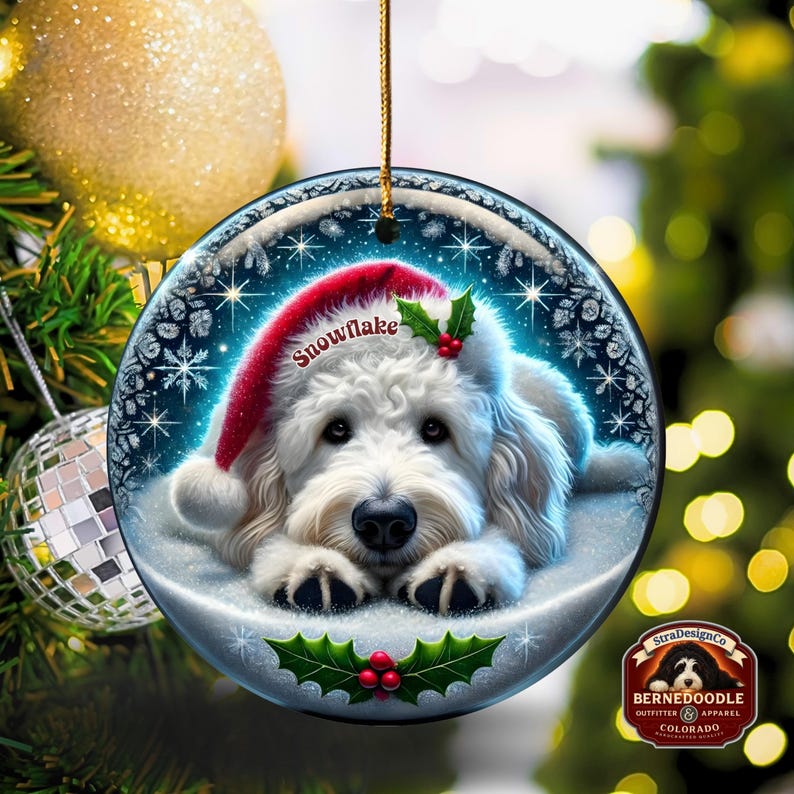 May include: A round Christmas ornament featuring a white Bernedoodle dog wearing a red Santa hat with the word "Snowflake". The ornament has a snowy background with holly and berries. The ornament is hanging from a gold string.