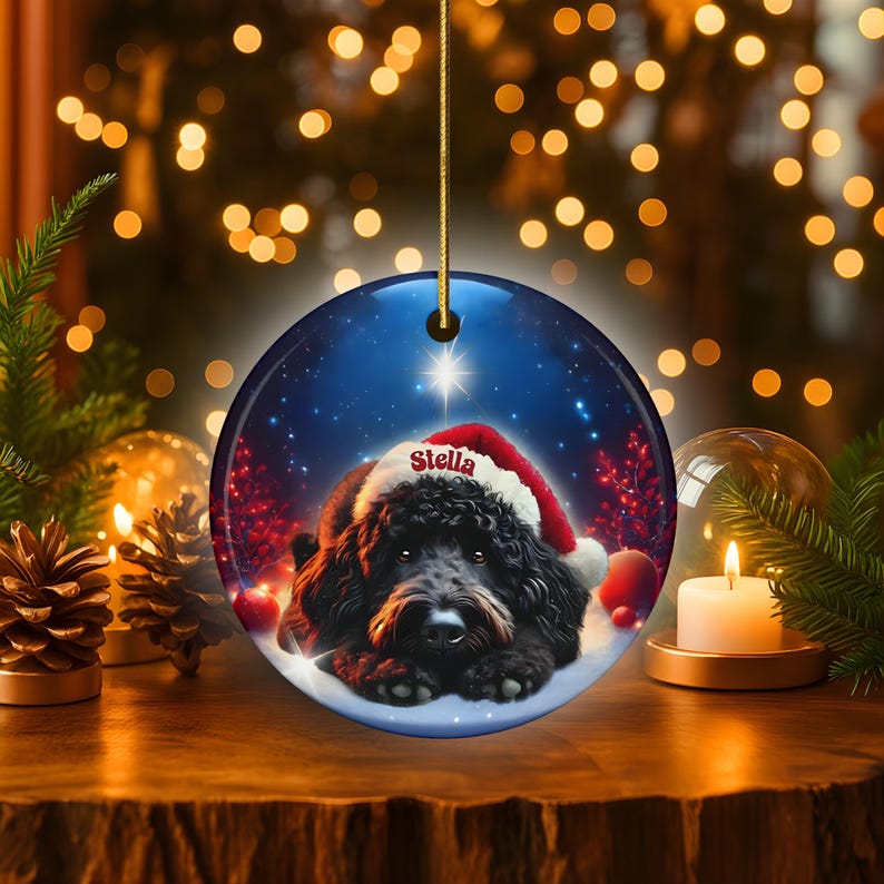 May include: Round Christmas ornament featuring a black dog wearing a Santa hat with the name "Stella". The ornament has a blue background with a star and snow effect. A gold string is attached for hanging.