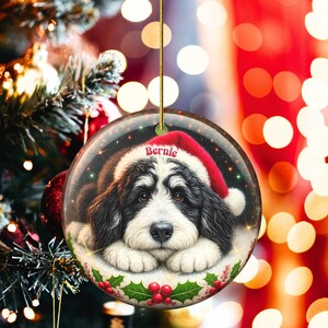 May include: A round Christmas ornament featuring a black and white dog wearing a Santa hat. The dog's name, "Bernie," is printed on the hat. The ornament is decorated with holly and red berries, and is hanging from a gold string.