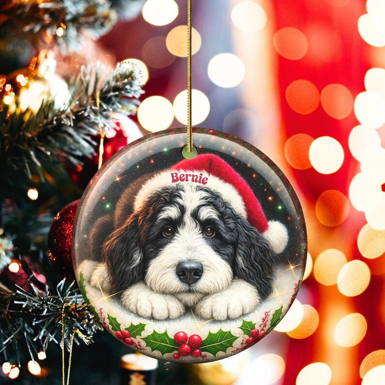 May include: A round Christmas ornament featuring a black and white dog wearing a Santa hat. The dog's name, "Bernie," is printed on the hat. The ornament is decorated with holly and red berries, and is hanging from a gold string.