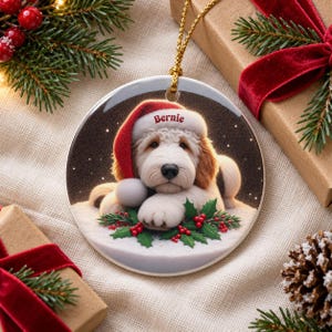 May include: A round ceramic Christmas ornament featuring a fluffy white and tan dog wearing a red Santa hat with the name Bernie printed on the brim. The dog is resting on a snowy surface decorated with green holly leaves and red berries.