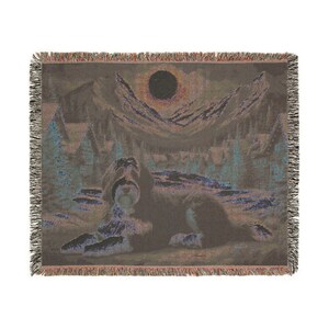 May include: Woven throw blanket featuring a dog lying down in a landscape with mountains, trees, and a dark sun. The color palette includes shades of brown, blue, and purple. The blanket has a fringed edge.