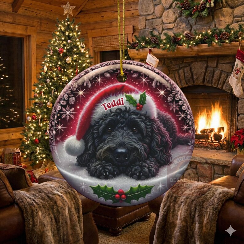 May include: A round Christmas ornament featuring a black dog wearing a Santa hat with the name "Teddi" on it. The ornament has a red and white border with snowflakes and holly berries. The background shows a cozy living room with a Christmas tree and a fireplace.