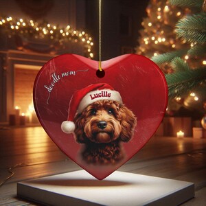 May include: Red heart-shaped Christmas ornament featuring a brown Doodle dog wearing a Santa hat with the name "Lucille." The ornament has the words "doodle mom" in script. A gold string is attached for hanging.