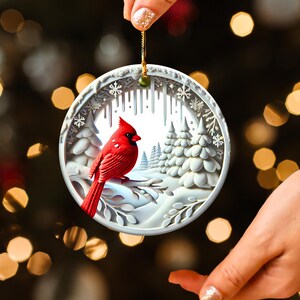 Peut inclure: Ornement de No&euml;l rond et blanc repr&eacute;sentant un cardinal rouge perch&eacute; sur une branche enneig&eacute;e. L'ornement pr&eacute;sente une sc&egrave;ne hivernale avec des arbres blancs et des flocons de neige, suspendu par une ficelle dor&eacute;e.