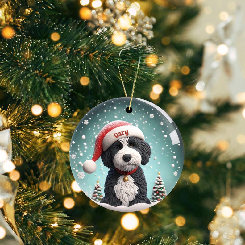 May include: Circular Christmas bauble depicting a black and white dog in a Santa hat, with the name "Gary". The bauble has a gold hanging string and is set against a snowy scene with two small Christmas trees.