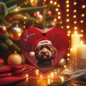 May include: A heart-shaped Christmas ornament with a red background and a portrait of a dog wearing a Santa hat. The dog's name, "Lucille," is printed on the hat. The words "doodle mom" are written on the ornament.