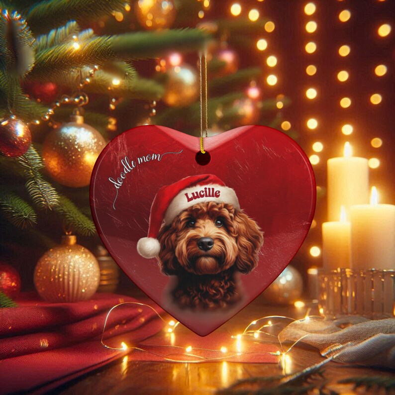 May include: A heart-shaped Christmas ornament with a red background and a portrait of a dog wearing a Santa hat. The dog's name, "Lucille," is printed on the hat. The words "doodle mom" are written on the ornament.