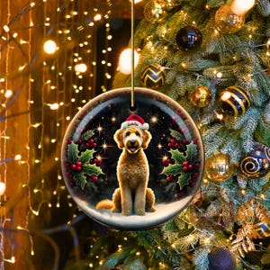 May include: A round Christmas ornament featuring a golden doodle dog wearing a Santa hat with the name "Goldie". The ornament is surrounded by holly and berries, set against a starry night background. The ornament hangs from a gold string.