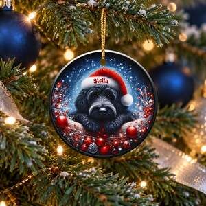 May include: A round ceramic Christmas ornament featuring a black dog wearing a red Santa hat with the name Stella printed in white. The dog is surrounded by red berries, small red ornaments, and a snowy blue background, hanging by a gold cord.