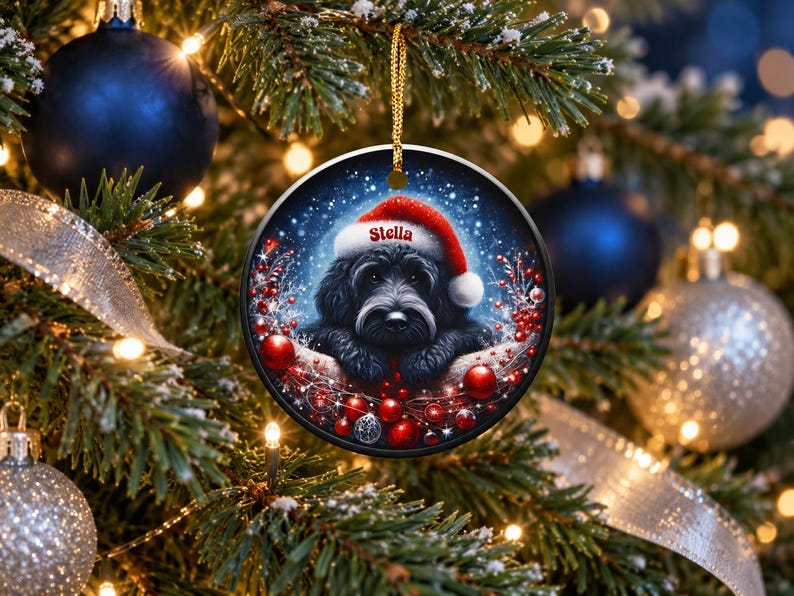 May include: A round ceramic Christmas ornament featuring a black dog wearing a red Santa hat with the name Stella printed in white. The dog is surrounded by red berries, small red ornaments, and a snowy blue background, hanging by a gold cord.
