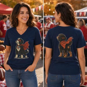 May include: A navy blue V-neck t-shirt featuring a graphic of a black and tan dog wearing a red superhero cape and a red eye mask. The front graphic shows the dog with a heart-shaped American flag patch, while the back shows the dog from behind.