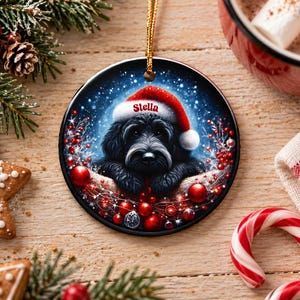 May include: A round ceramic Christmas ornament featuring a black dog wearing a red Santa hat with the name Stella printed on the white trim. The dog is surrounded by red ornaments, berries, and a snowy blue background, hanging by a gold cord.