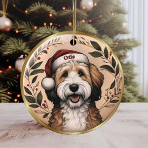 May include: A round Christmas ornament featuring a smiling dog wearing a Santa hat with the name "Otis". The dog is surrounded by a green and red leaf design, with a gold border and hanging loop.