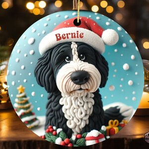 Puede incluir: Adorno navideño redondo con un perro blanco y negro con un gorro de Papá Noel con el nombre "Bernie". El fondo es azul con copos de nieve blancos y un pequeño árbol de Navidad. Se adjunta una cuerda dorada para colgar.