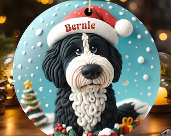 May include: Round Christmas ornament featuring a black and white dog wearing a Santa hat with the name "Bernie" on it. The ornament has a blue background with white snowflakes and a small Christmas tree. A gold string is attached for hanging.