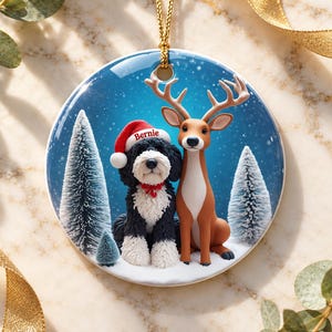 May include: A round ceramic ornament featuring a black and white dog wearing a red Santa hat with the name Bernie printed on it, sitting next to a brown deer in a snowy forest with pine trees under a blue sky, suspended by a gold cord.