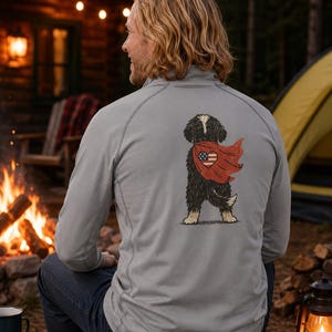 K&ouml;nnte beinhalten: Ein hellgrauer Pullover mit langen &Auml;rmeln und einem R&uuml;ckendruck, der einen schwarz-wei&szlig;en Hund mit einem roten Umhang zeigt. Auf dem Umhang befindet sich ein herzf&ouml;rmiges Emblem mit der amerikanischen Flagge. Der Hund ist von hinten dargestellt.
