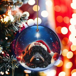 May include: A round Christmas ornament featuring a black dog wearing a Santa hat with the name "Stella" on it. The ornament has a blue background with a star and a snowy ground. The ornament is hanging from a gold string.