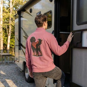 Pu&ograve; includere: Una felpa con zip a un quarto di colore rosa polveroso con una grafica di un cane nero con un mantello rosso. La felpa &egrave; realizzata in un materiale morbido e viene indossata da una persona vicino a un camper.