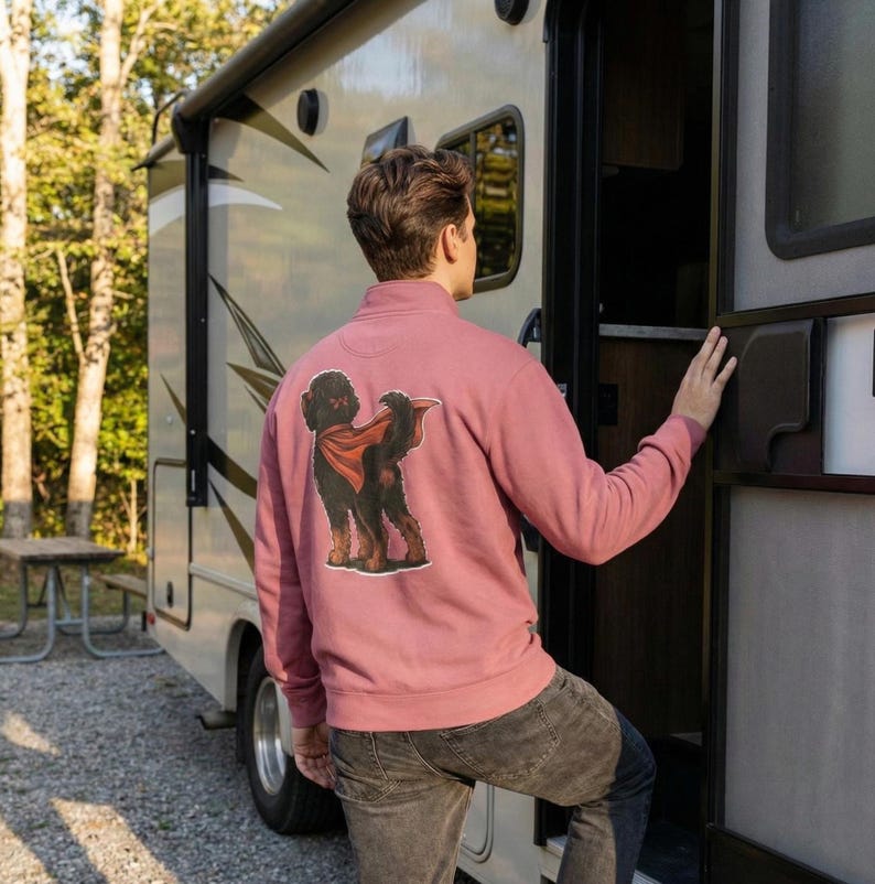 Pu&ograve; includere: Una felpa con zip a un quarto di colore rosa polveroso con una grafica di un cane nero con un mantello rosso. La felpa &egrave; realizzata in un materiale morbido e viene indossata da una persona vicino a un camper.