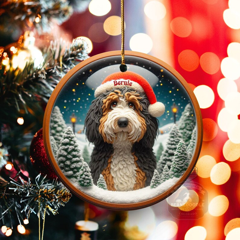 Op de afbeelding: Een ronde kerstversiering met een Bernedoodle-hond met een kerstmuts met de naam "Bernie". De versiering heeft een winterse achtergrond met besneeuwde bomen en een sterrenhemel. De versiering is omlijst met hout.