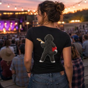 May include: A black short-sleeved t-shirt featuring a graphic on the back of a fluffy black dog standing on its hind legs and playing a light-colored electric guitar with a red heart-shaped body, paired with blue denim jeans.