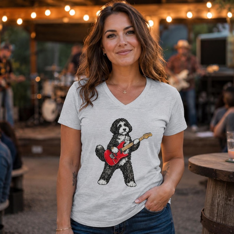 May include: A light gray V-neck t-shirt featuring a graphic illustration of a black and white dog standing on its hind legs while playing a bright red electric guitar. The dog has shaggy fur and is positioned in the center of the shirt.