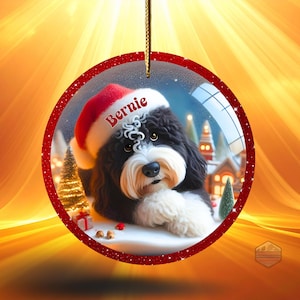 K&ouml;nnte beinhalten: Runder Weihnachtsschmuck mit einem schwarz-wei&szlig;en Hund, der einen Weihnachtsmannhut mit dem Namen "Bernie" tr&auml;gt. Der Schmuck hat einen roten Glitzerrahmen und eine goldene Schnur zum Aufh&auml;ngen. Im Inneren ist eine Winterszene zu sehen.