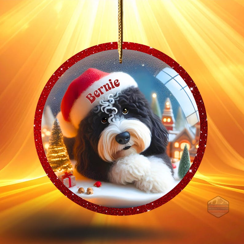 K&ouml;nnte beinhalten: Runder Weihnachtsschmuck mit einem schwarz-wei&szlig;en Hund, der einen Weihnachtsmannhut mit dem Namen "Bernie" tr&auml;gt. Der Schmuck hat einen roten Glitzerrahmen und eine goldene Schnur zum Aufh&auml;ngen. Im Inneren ist eine Winterszene zu sehen.